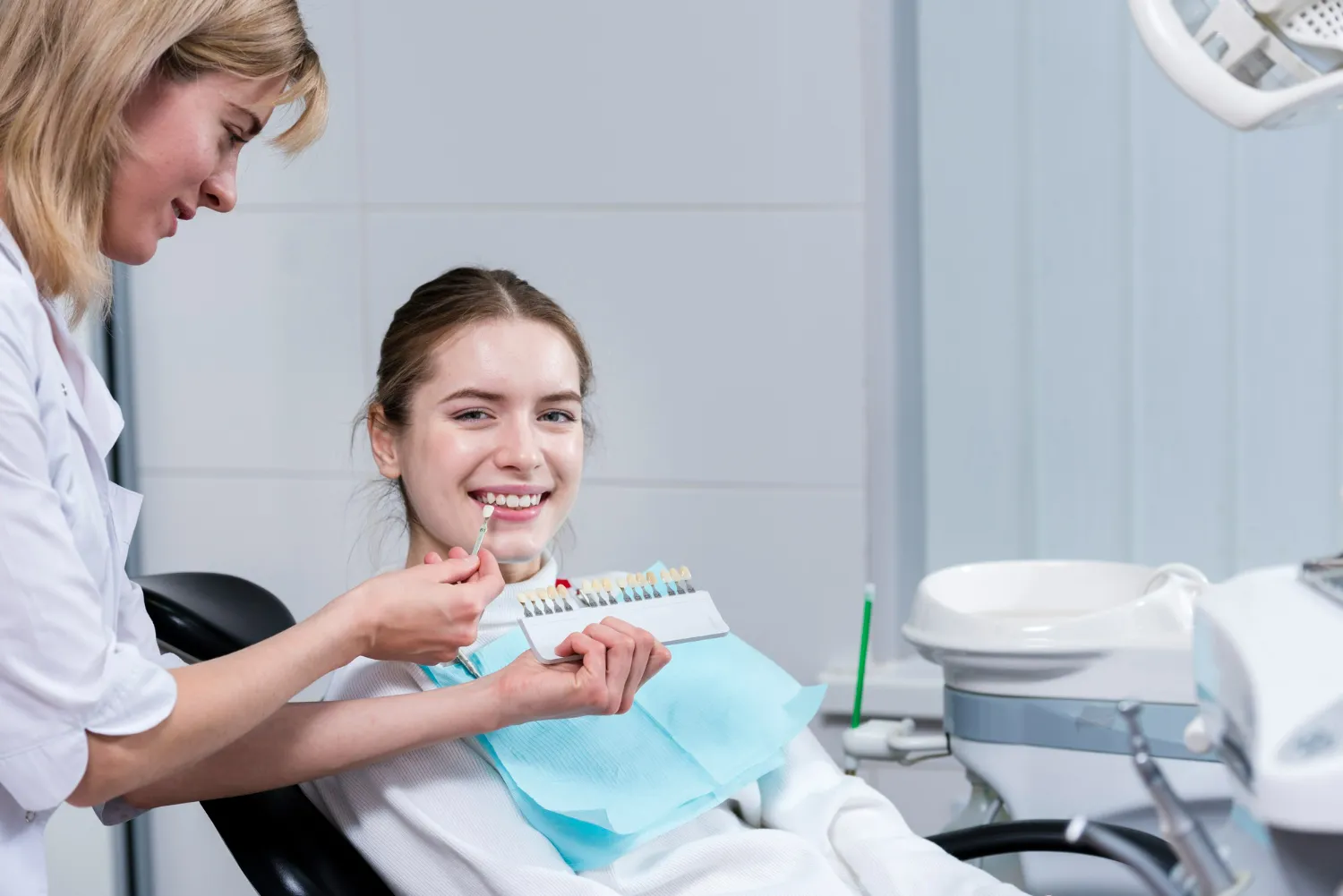 Dentist holding a tooth shade guide and discussing whitening options with a smiling patient in the chair.