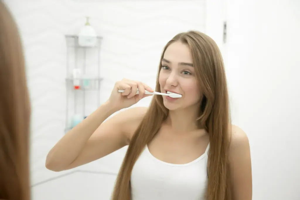Young woman brushing her teeth at a bathroom mirror as part of a daily oral hygiene routine to protect porcelain veneers.